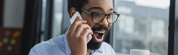 Hombre de negocios afroamericano asombrado hablando por teléfono móvil y sosteniendo la taza en la oficina, pancarta - foto de stock