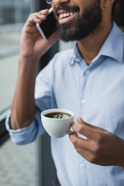 Ausgeschnittene Ansicht einer Tasse Kaffee in der Hand eines verschwommenen afrikanisch-amerikanischen Geschäftsmannes, der auf dem Smartphone spricht — Stockfoto
