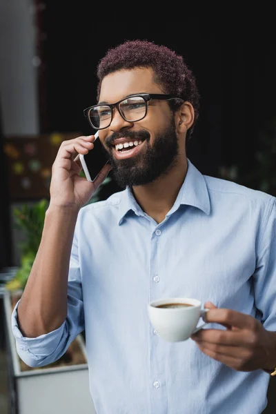 Joven hombre de negocios afroamericano sosteniendo una taza de café y hablando en el teléfono inteligente en la oficina - foto de stock