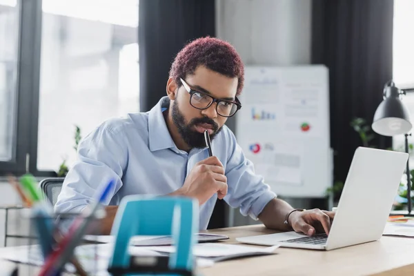 Pensive gestionnaire afro-américain tenant stylo et regardant ordinateur portable dans le bureau — Photo de stock