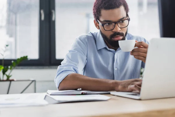 Африканський американський бізнесмен в окулярах тримає чашку кави і за допомогою ноутбука біля записника і паперів в офісі — стокове фото