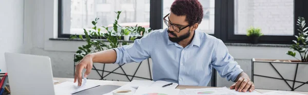 Afrikanischer Geschäftsmann arbeitet mit Dokumenten in der Nähe von Laptop auf Tisch im Büro, Banner — Stockfoto