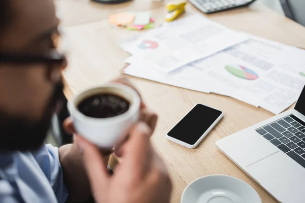 Dispositivi e carte vicino sfocato uomo d'affari afroamericano in possesso di caffè in ufficio — Foto stock