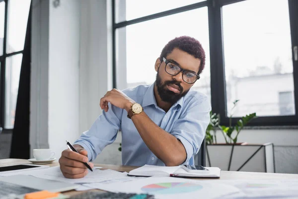 Африканський американський менеджер, який тримає ручку біля паперів і гаджетів в офісі — стокове фото