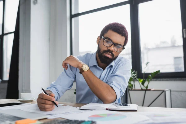 Gestionnaire afro-américain tenant stylo près des papiers et gadgets au bureau — Photo de stock