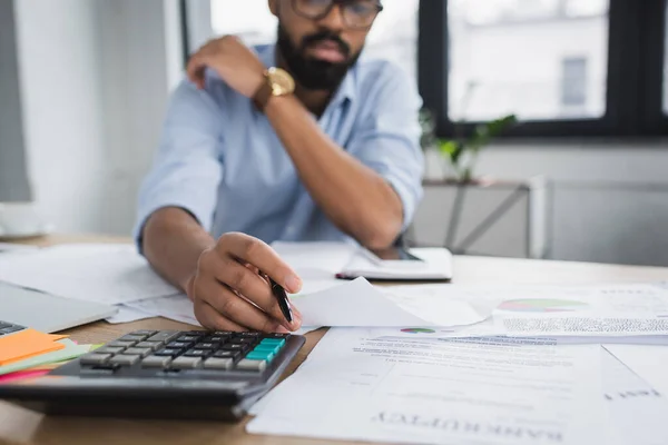 Homme d'affaires afro-américain flou tenant un stylo près des papiers et une calculatrice sur la table — Photo de stock