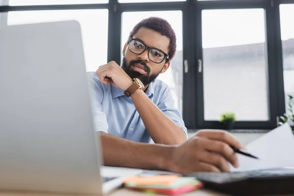 Африканський американський бізнесмен, який дивиться на ноутбук біля розмитого документа в офісі — стокове фото