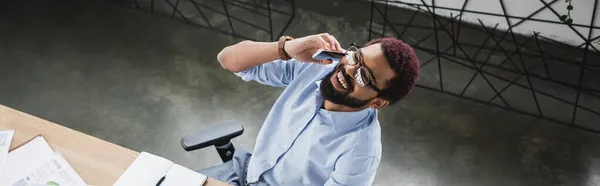 Вид веселого африканского бизнесмена в очках, говорящего по смартфону рядом с рабочим столом в офисе, баннер — стоковое фото