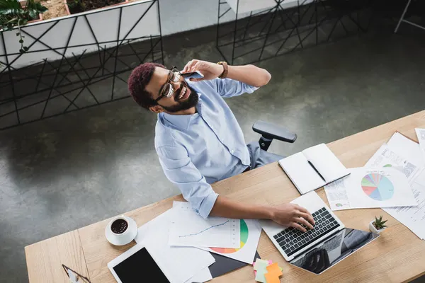 Positiver afrikanisch-amerikanischer Geschäftsmann im Gespräch mit Smartphone in der Nähe von Gadgets, Kaffee und Dokumenten im Büro — Stockfoto