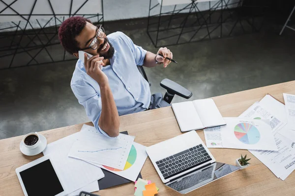 Vista aerea dell'uomo d'affari afroamericano che parla sul cellulare vicino a documenti, caffè e gadget in ufficio — Foto stock