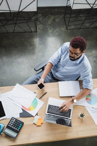 Overhead-Ansicht eines afrikanisch-amerikanischen Geschäftsmannes mit Smartphone und Laptop in der Nähe von Dokumenten im Büro — Stockfoto