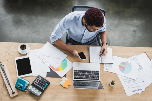 Overhead view of african american businessman writing on notebook near gadgets and papers on table — Stock Photo