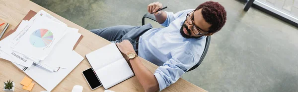 Overhead view of african american businessman holding pen near notebook, papers and smartphone, banner — Stock Photo