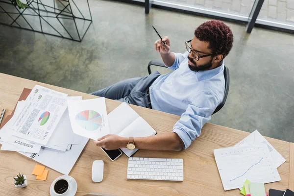 Overhead-Ansicht eines afrikanisch-amerikanischen Geschäftsmannes in Brille mit Dokument in der Nähe von Kaffee und Smartphone auf dem Arbeitstisch — Stockfoto