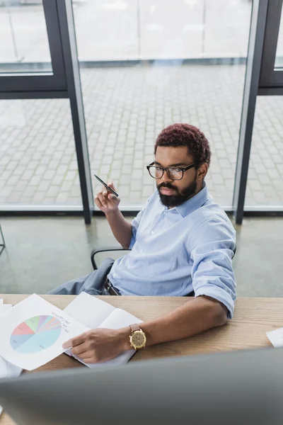 African american businessman holding pen and document near blurred computer monitor in office — Stock Photo