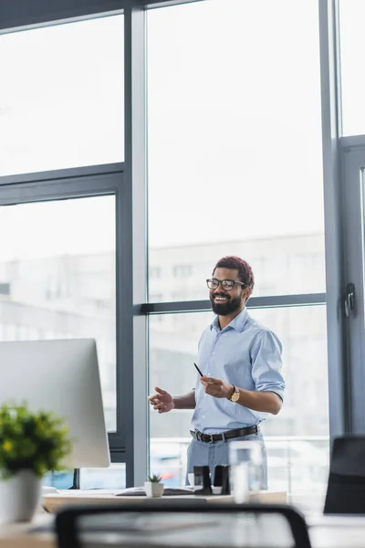 Joyful african american businessman holding pen near computer on table in office — Stock Photo