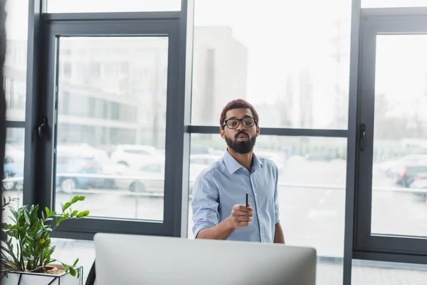 Homme d'affaires afro-américain parlant pendant le chat vidéo et tenant un stylo près de l'ordinateur au bureau — Photo de stock