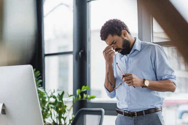 Empresário afro-americano exausto segurando óculos e tocando os olhos perto do computador no escritório — Fotografia de Stock