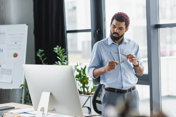 Jovem gerente afro-americano segurando óculos perto do monitor do computador e flipchart borrado no escritório — Fotografia de Stock