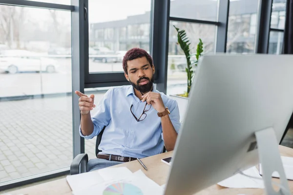 Homme d'affaires afro-américain tenant des lunettes et pointant du doigt près de l'écran d'ordinateur au bureau — Photo de stock