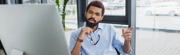 Gerente afroamericano señalando con el dedo y sosteniendo las gafas cerca del monitor de la computadora, pancarta - foto de stock