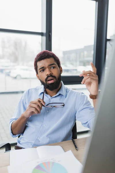 Empresário afro-americano segurando óculos e gesticulando perto de computador borrado no escritório — Fotografia de Stock