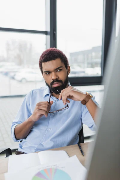 Young african american businessman holding eyeglasses near notebook and computer monitor — Stock Photo