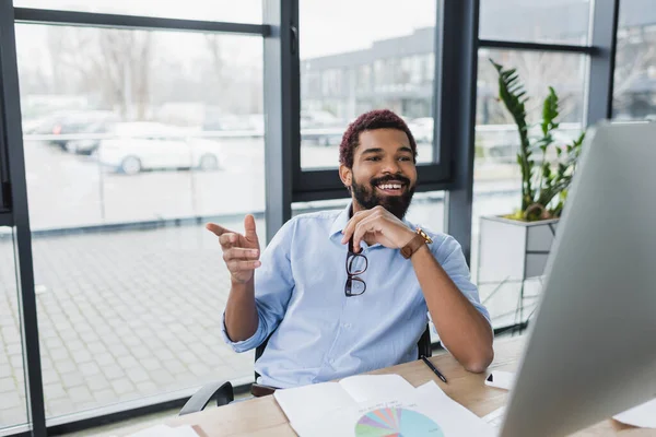 Empresario afroamericano positivo sosteniendo anteojos y señalando con el dedo cerca de la computadora en la oficina - foto de stock