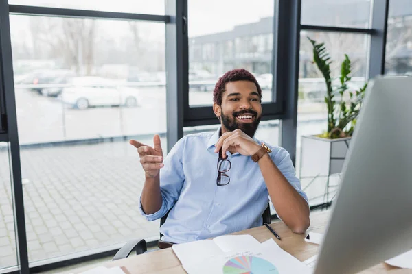 Positivo uomo d'affari afroamericano che tiene gli occhiali e punta con il dito vicino al computer in ufficio — Foto stock