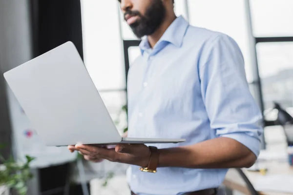 Vista recortada del hombre de negocios afroamericano en camisa que sostiene el ordenador portátil en la oficina - foto de stock