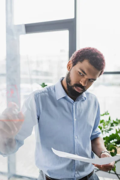 African american businessman with paper writing on glass board in office — Stock Photo