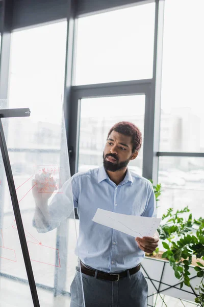 African american businessman with document writing on glass board in office — Stock Photo