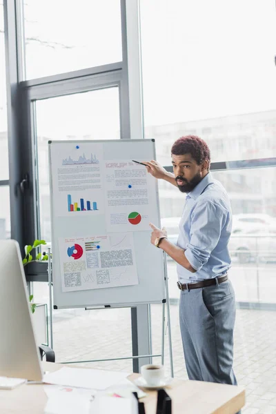 Hombre de negocios afroamericano apuntando al rotafolio con gráficos cerca de la computadora durante una videollamada en la oficina - foto de stock