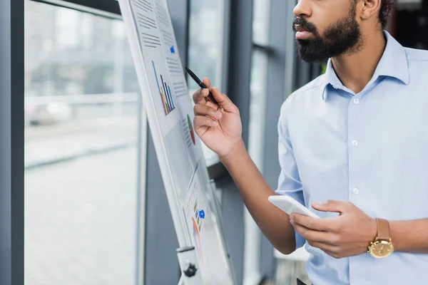Cropped view of african american businessman with pen and smartphone standing near flip chart in office — Stock Photo