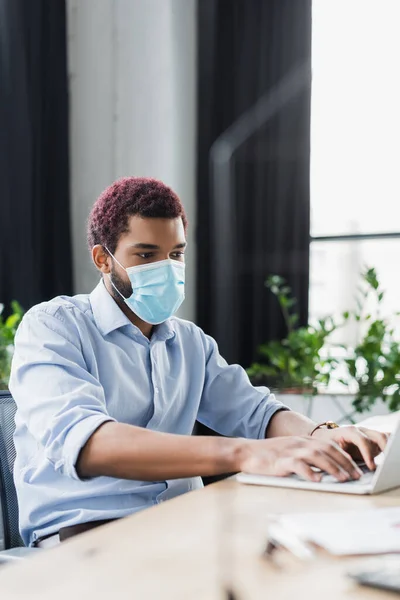 Uomo d'affari afroamericano in maschera medica utilizzando il computer portatile in ufficio — Foto stock