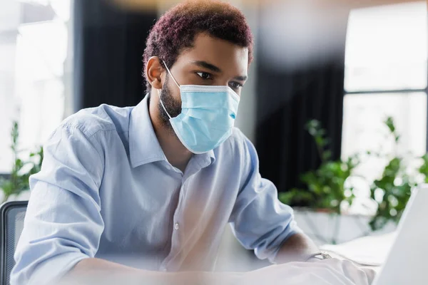 Молодой африканский бизнесмен в медицинской маске смотрит на размытый ноутбук — стоковое фото