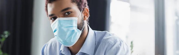 Hombre de negocios afroamericano en máscara médica mirando a la cámara en la oficina, pancarta - foto de stock