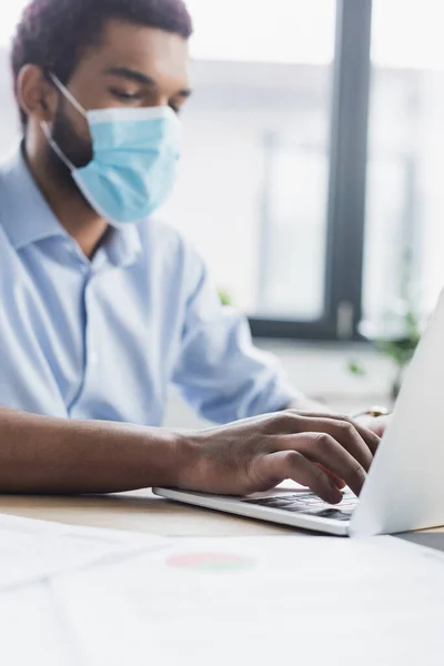 Blurred african american businessman in medical mask using laptop in office — Stock Photo