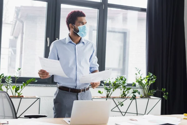 Hombre de negocios afroamericano con máscara médica sosteniendo papeles cerca de la computadora portátil en la oficina - foto de stock