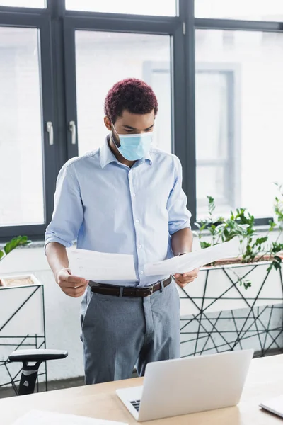 Homme d'affaires afro-américain en masque médical et vêtements formels à la recherche de documents près d'un ordinateur portable au bureau — Photo de stock