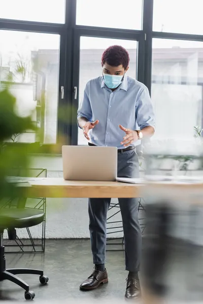 Young african american businessman in medical mask having video chat on laptop in office — Stock Photo
