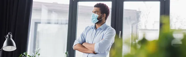 Hombre de negocios afroamericano con máscara médica de pie con los brazos cruzados en la oficina, pancarta - foto de stock