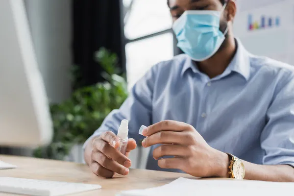 Verschwommener afrikanisch-amerikanischer Geschäftsmann in medizinischer Maske hält Händedesinfektionsmittel in der Nähe von Computer im Büro — Stockfoto