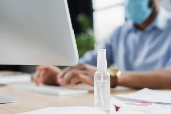 Desinfectante de manos cerca de la computadora y desenfocado hombre de negocios afroamericano en máscara médica - foto de stock