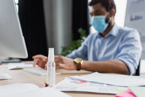 Disinfettante per le mani vicino sfocato uomo d'affari afroamericano in maschera medica utilizzando il computer in ufficio — Foto stock