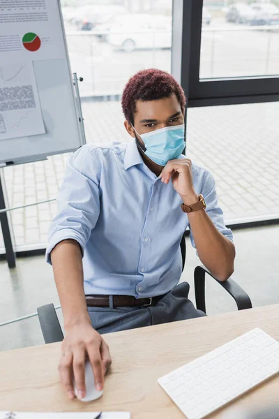 Africano americano gerente em máscara médica usando mouse computador perto de flip chart no escritório — Fotografia de Stock