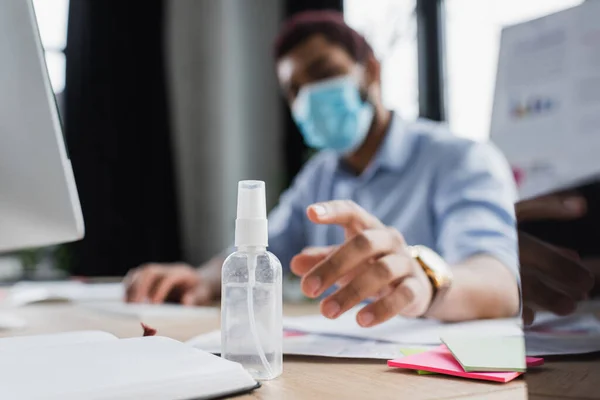 Blurred african american manager in protective mask taking hand sanitizer near computer and notebook in office — Stock Photo