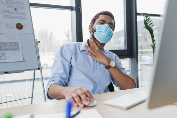 Sick african american businessman in medical mask touching neck while using laptop in office — Stock Photo