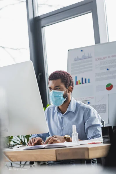 Empresario afroamericano en máscara médica usando portátil cerca de desinfectante de manos en la oficina - foto de stock