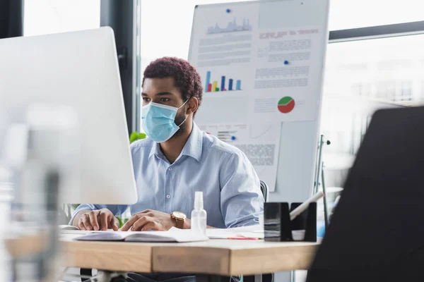 Африканский американский бизнесмен в защитной маске с помощью компьютера возле размытой флип-карты в офисе — стоковое фото
